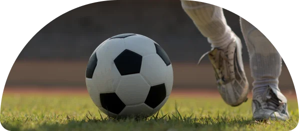 Gros plan sur un ballon de soccer sur l'herbe avec la jambe d'un joueur en mouvement, prête à frapper. La scène transmet anticipation et concentration, dans un stade flou.
