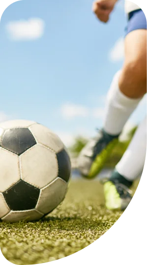 Gros plan sur un ballon de soccer sur un gazon vert alors qu'un joueur en mouvement, portant des chaussettes blanches et des crampons, se prépare à frapper. Journée ensoleillée et dynamique.