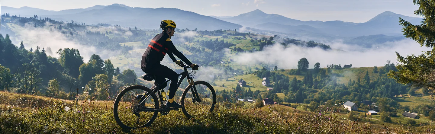 Un cycliste portant un casque jaune roule sur une colline herbeuse, surplombant une vallée brumeuse avec des maisons éparpillées. L'arrière-plan présente des montagnes luxuriantes et un ciel serein.