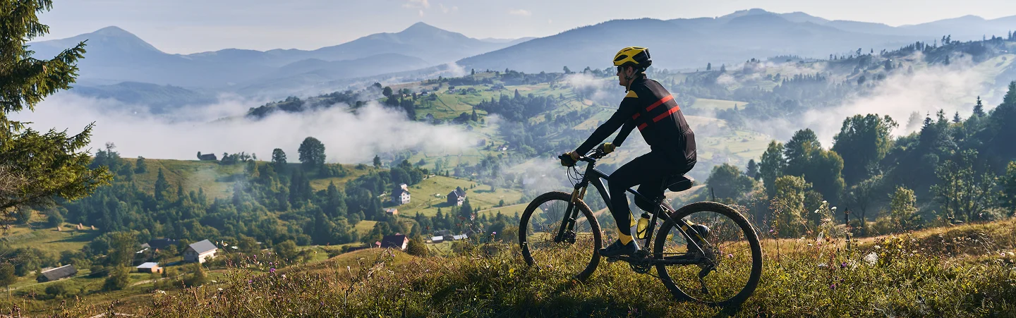 Un cycliste portant un casque jaune roule sur une colline herbeuse, surplombant une vallée brumeuse avec des maisons éparpillées. L'arrière-plan présente des montagnes luxuriantes et un ciel serein.